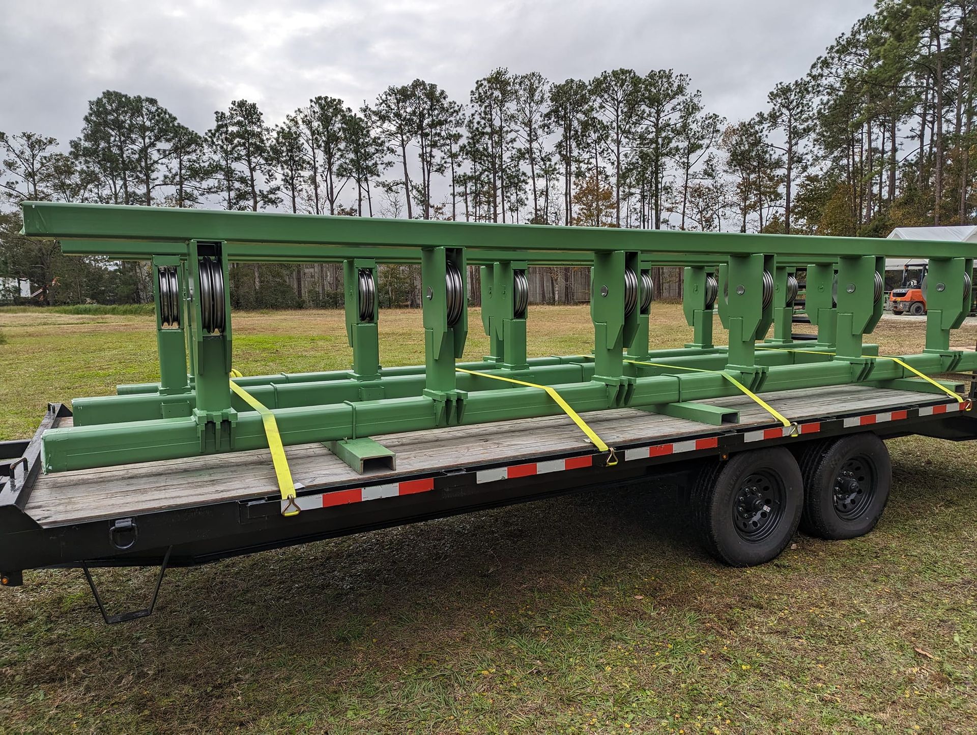 Green metal frame secured on a flatbed trailer with tie-down straps in a grassy outdoor setting.