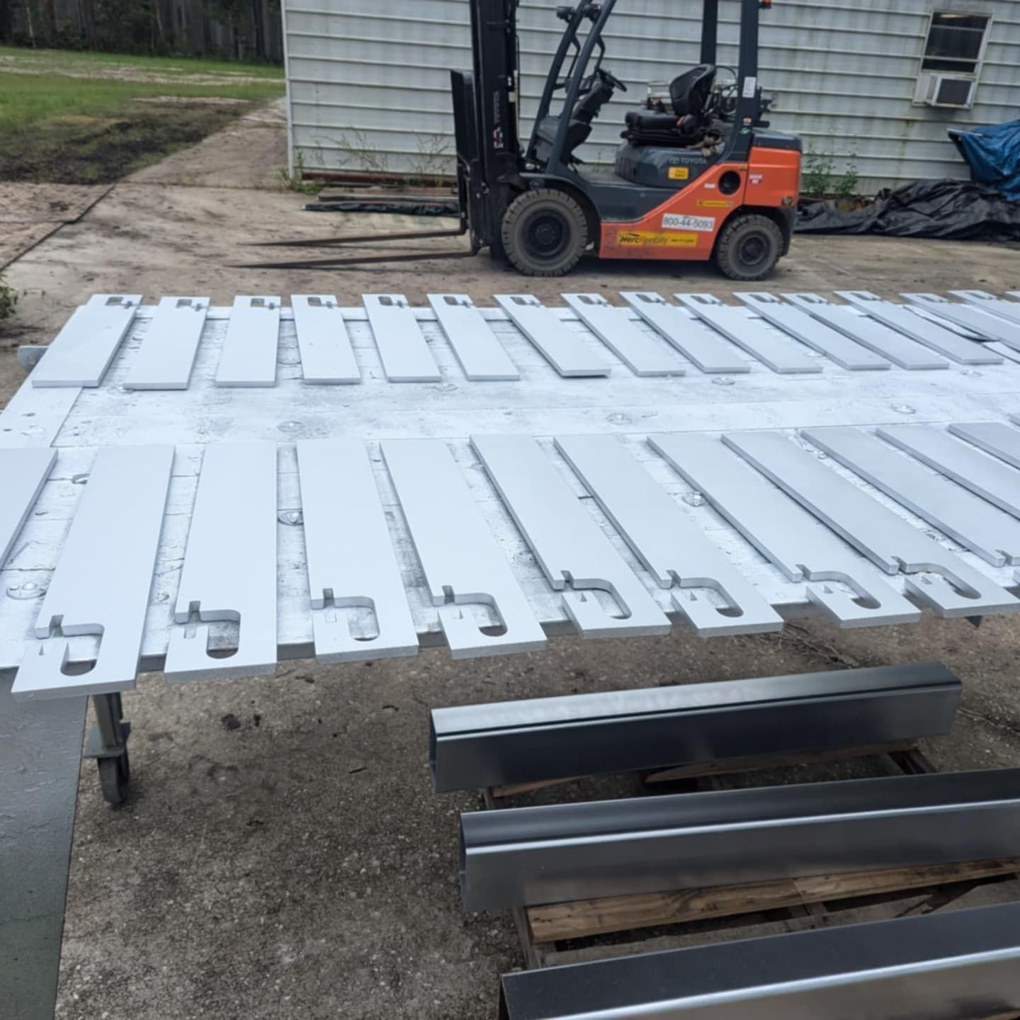 White painted metal parts laid out on a table with a forklift in the background, outside a building.