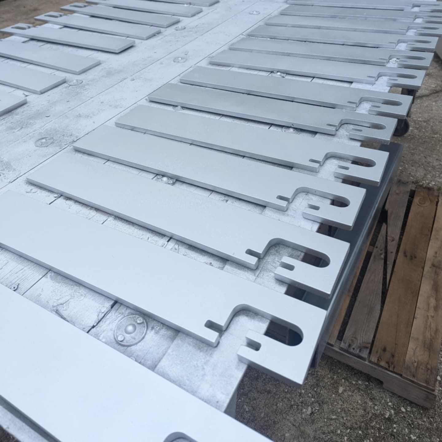 Painted, rectangular metal pieces with notched ends arranged on a wooden pallet for drying.