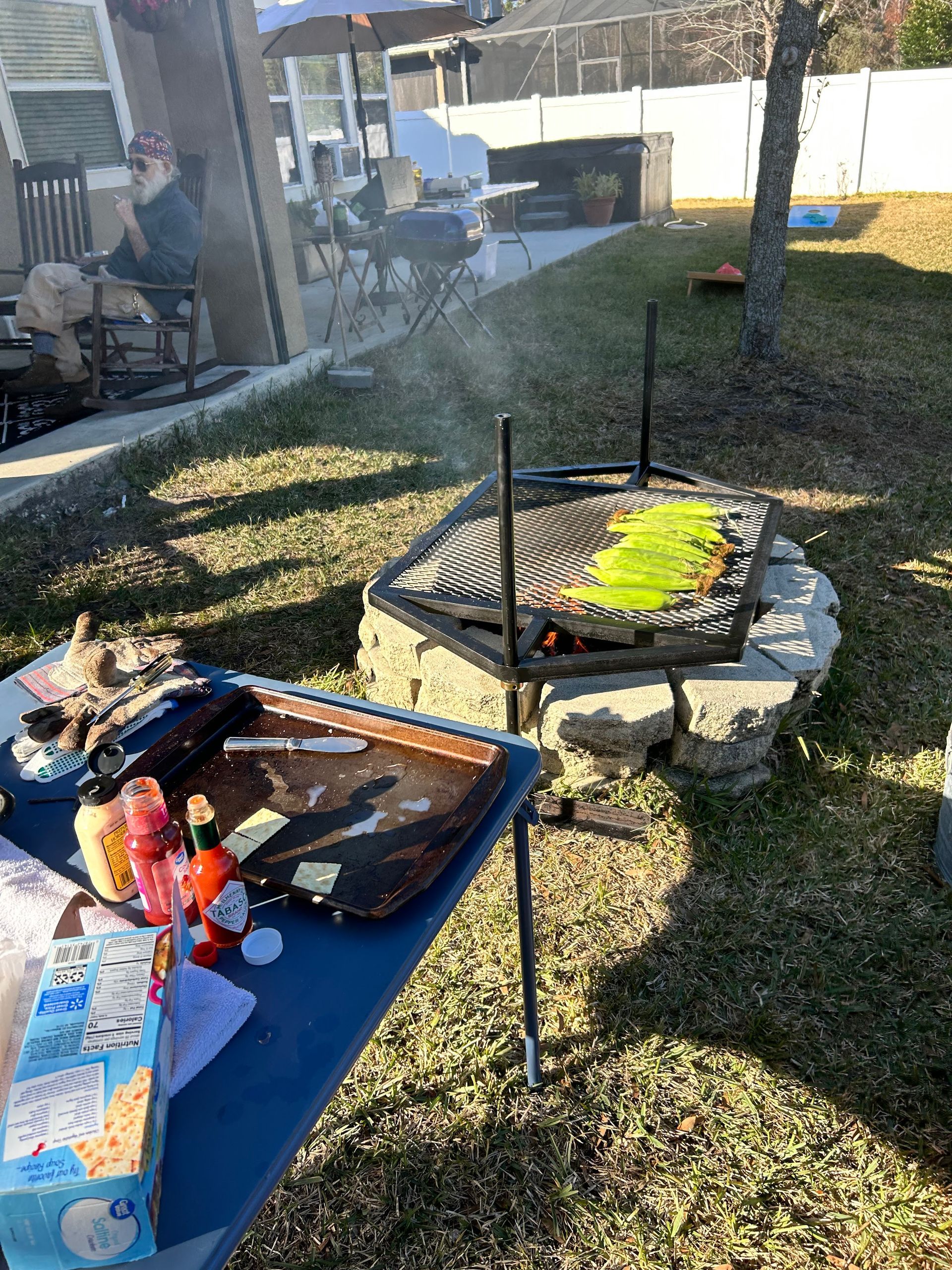 Grilling outdoors with a person sitting on a patio, fire pit cooking, grilling some green peppers, blue table, and lawn.