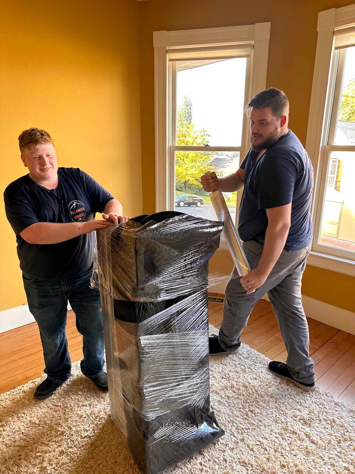 Two men are wrapping boxes in plastic wrap in a living room.