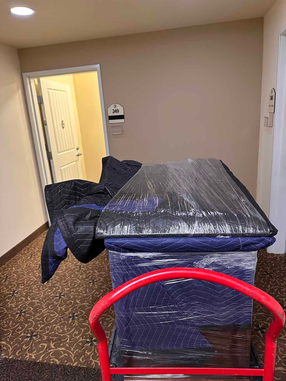 A dresser is wrapped in plastic and sitting on a dolly in a hallway.