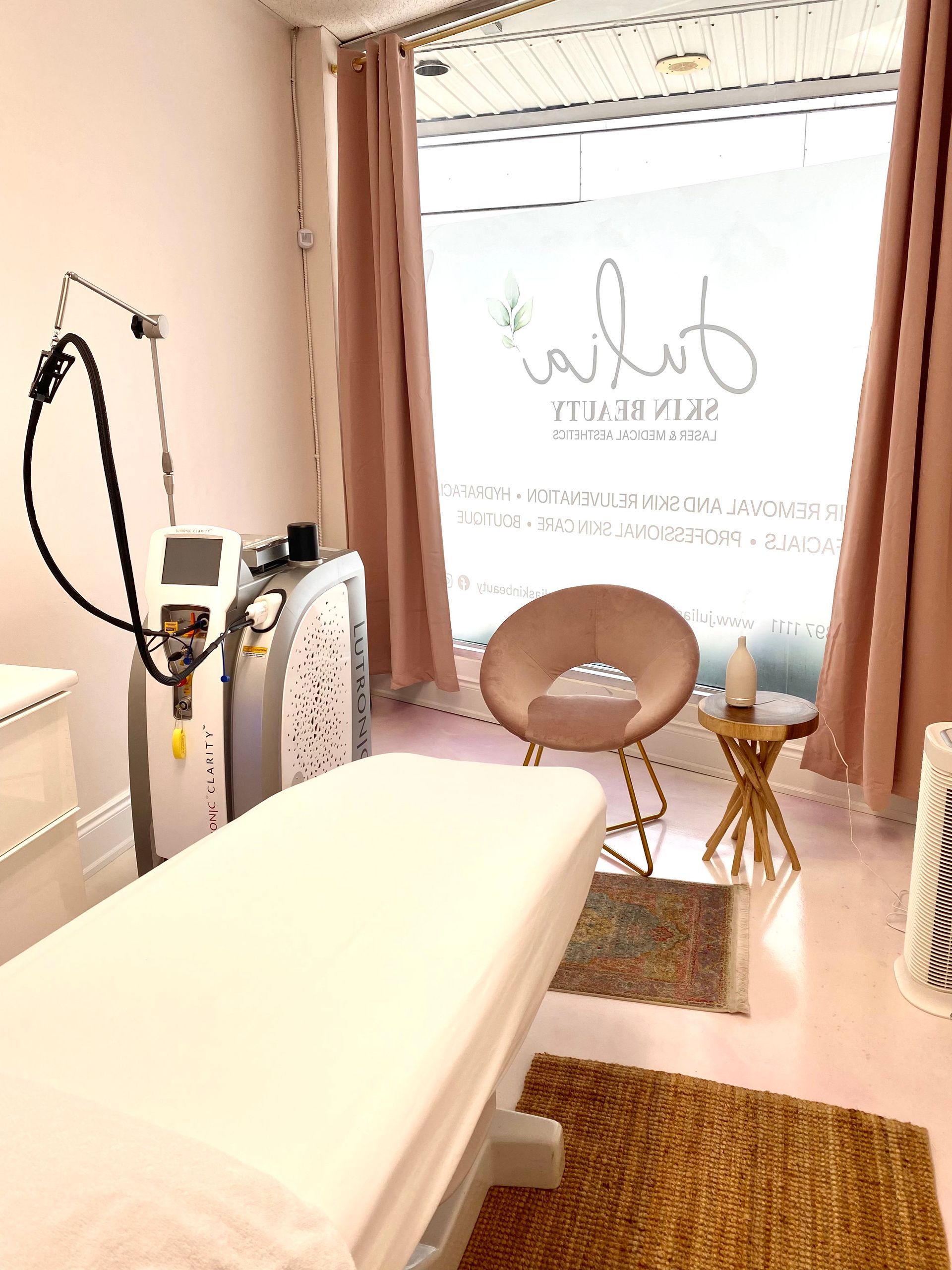 A clinical aesthetic room featuring a white treatment table, medical laser equipment, and a small pink chair by a window.