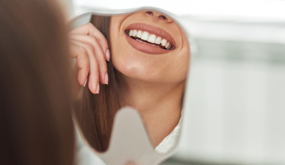 A person smiles at their reflection in a hand mirror, focusing on their teeth.