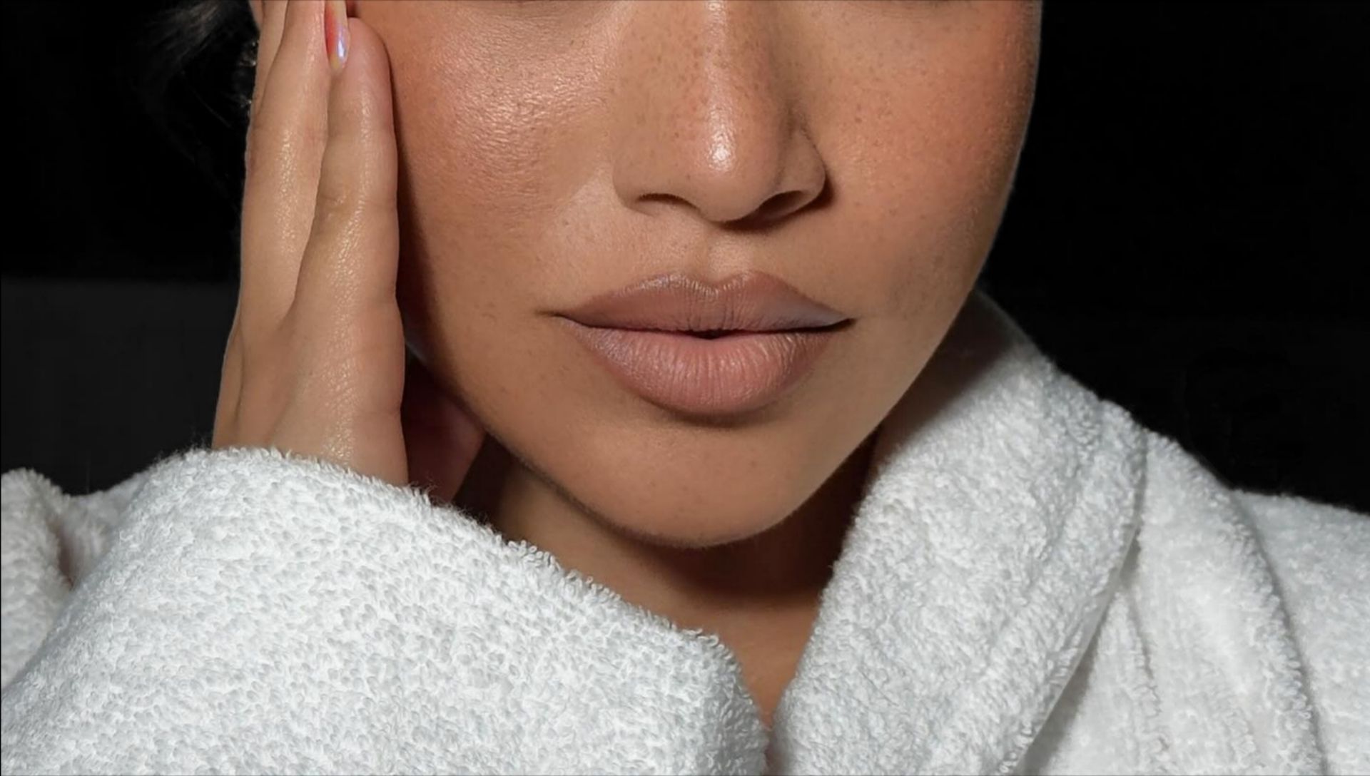 A close-up of a person's face wearing a white robe, with their hand gently touching their cheek against a dark background.