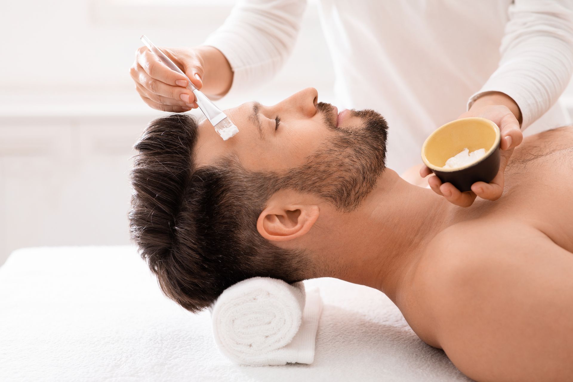 An aesthetician applies a white face mask with a brush to a man’s forehead while he relaxes on a spa treatment table.