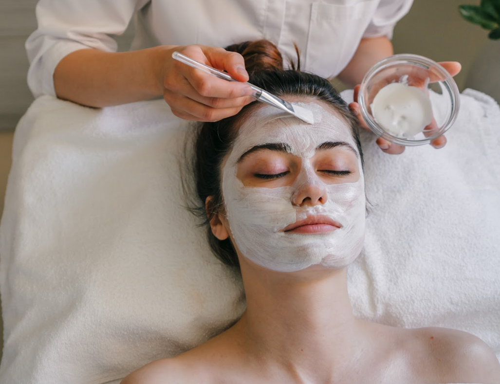 A person lies on a table while a professional applies a white facial mask with a brush from a small glass bowl.