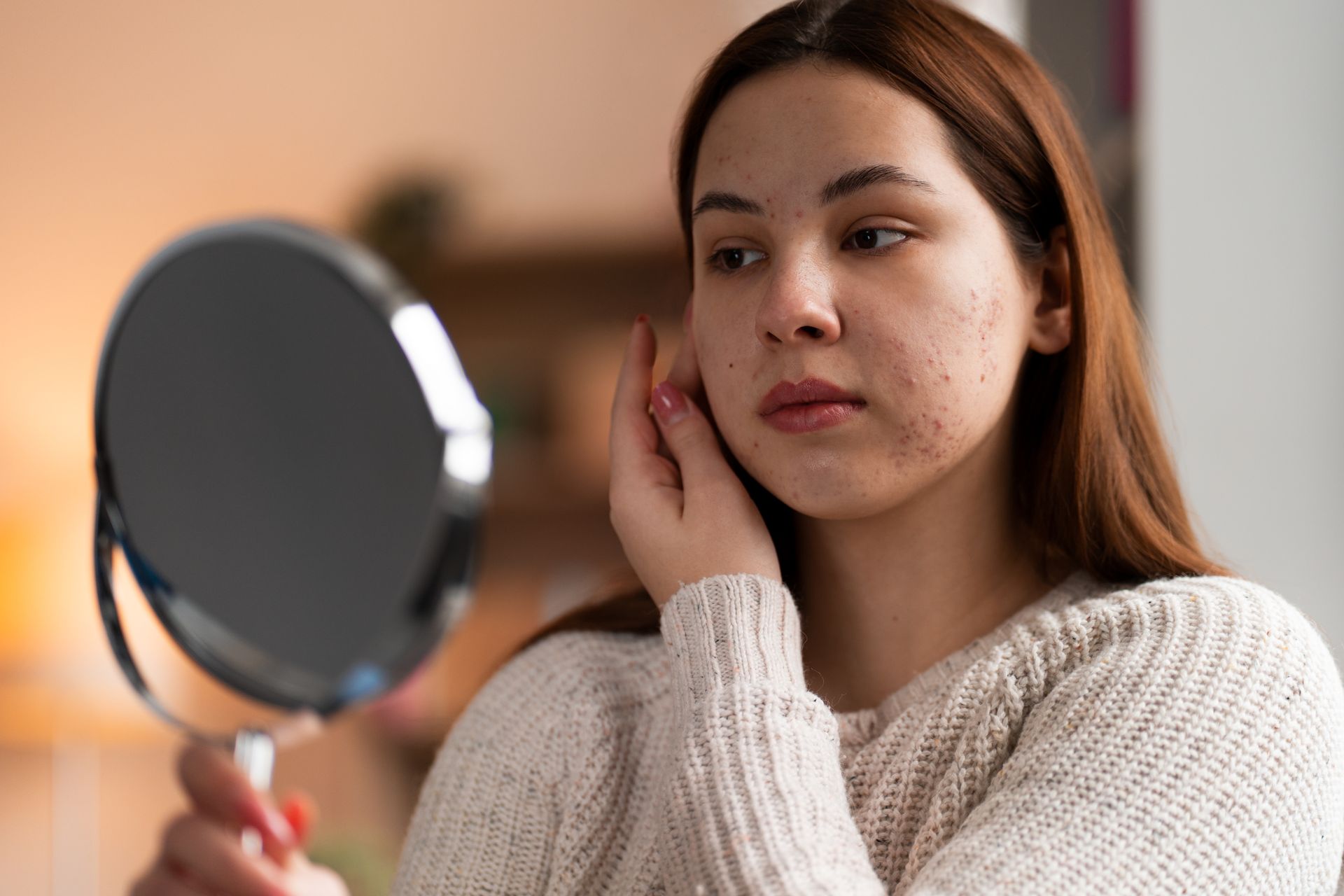 Woman looking at her face in a mirror, inspecting skin with acne; indoors.