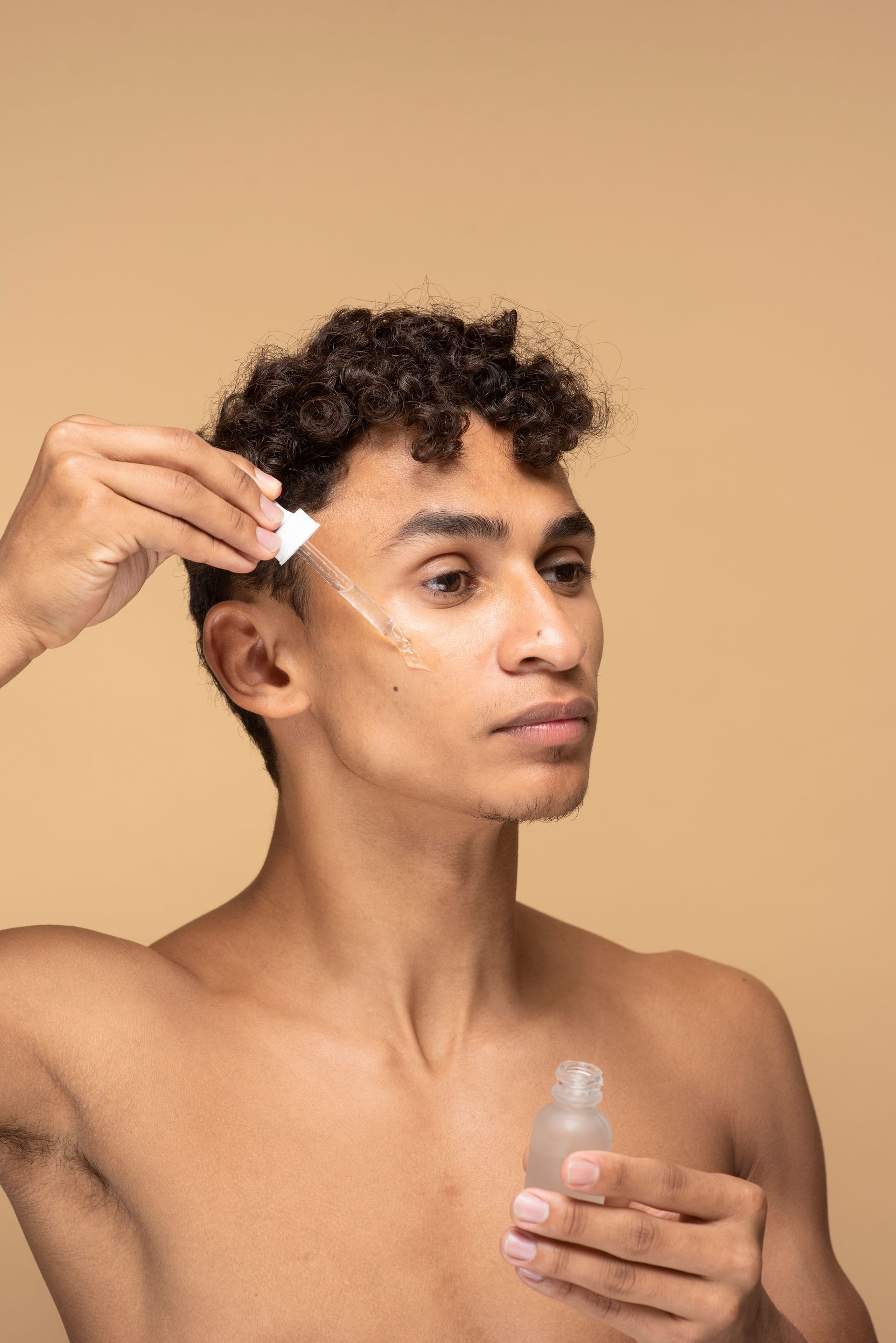 A shirtless man is applying a serum to his face.