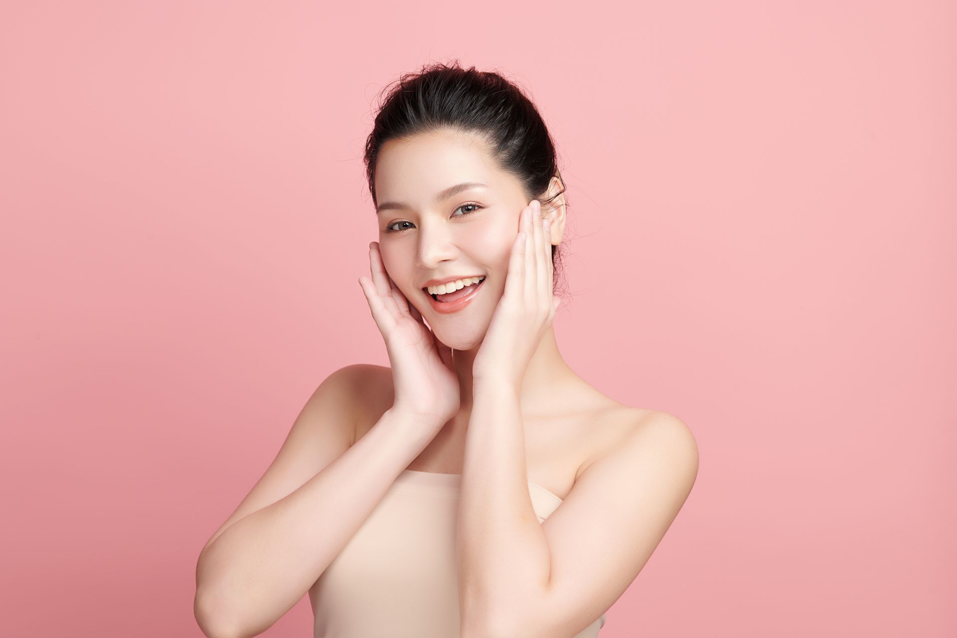 A woman is smiling and touching her face with her hands on a pink background.