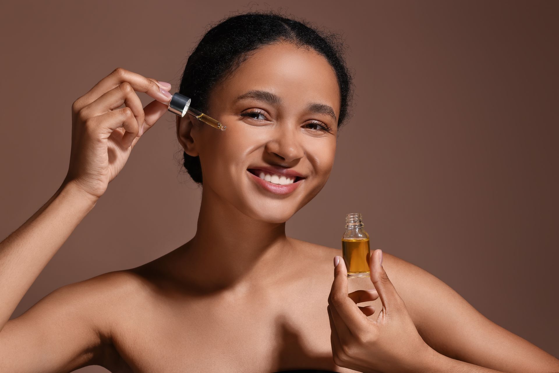 A woman is smiling while applying a serum to her face.