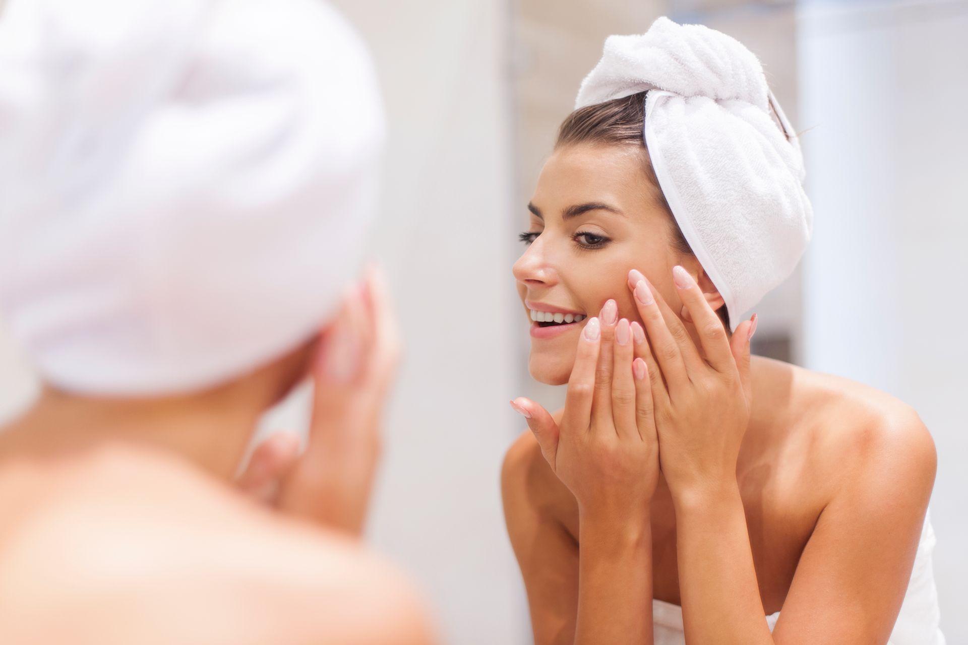 A woman with a towel wrapped around her head is looking at her face in the mirror.