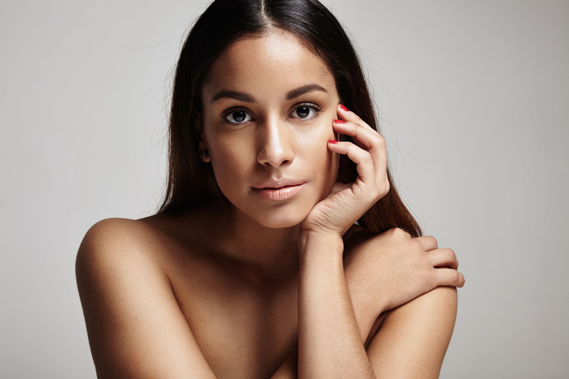 A person with long brown hair rests their chin on their hand, looking toward the camera against a gray background.