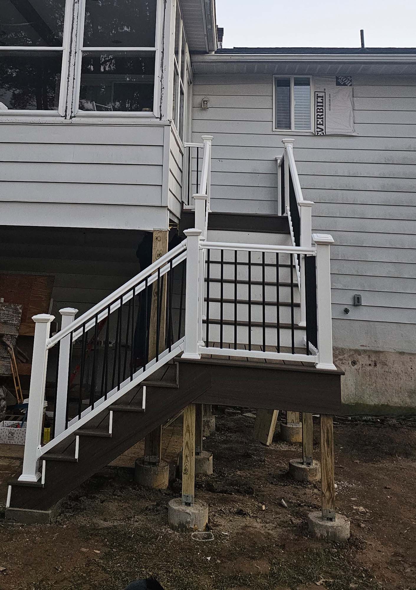 Staircase with white railing, black metal balusters, and brown steps leading to a house with white siding.