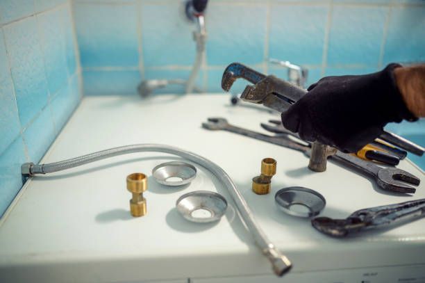 Plumber's hands wearing gloves holding wrench, tools and parts on a white surface in a bathroom.
