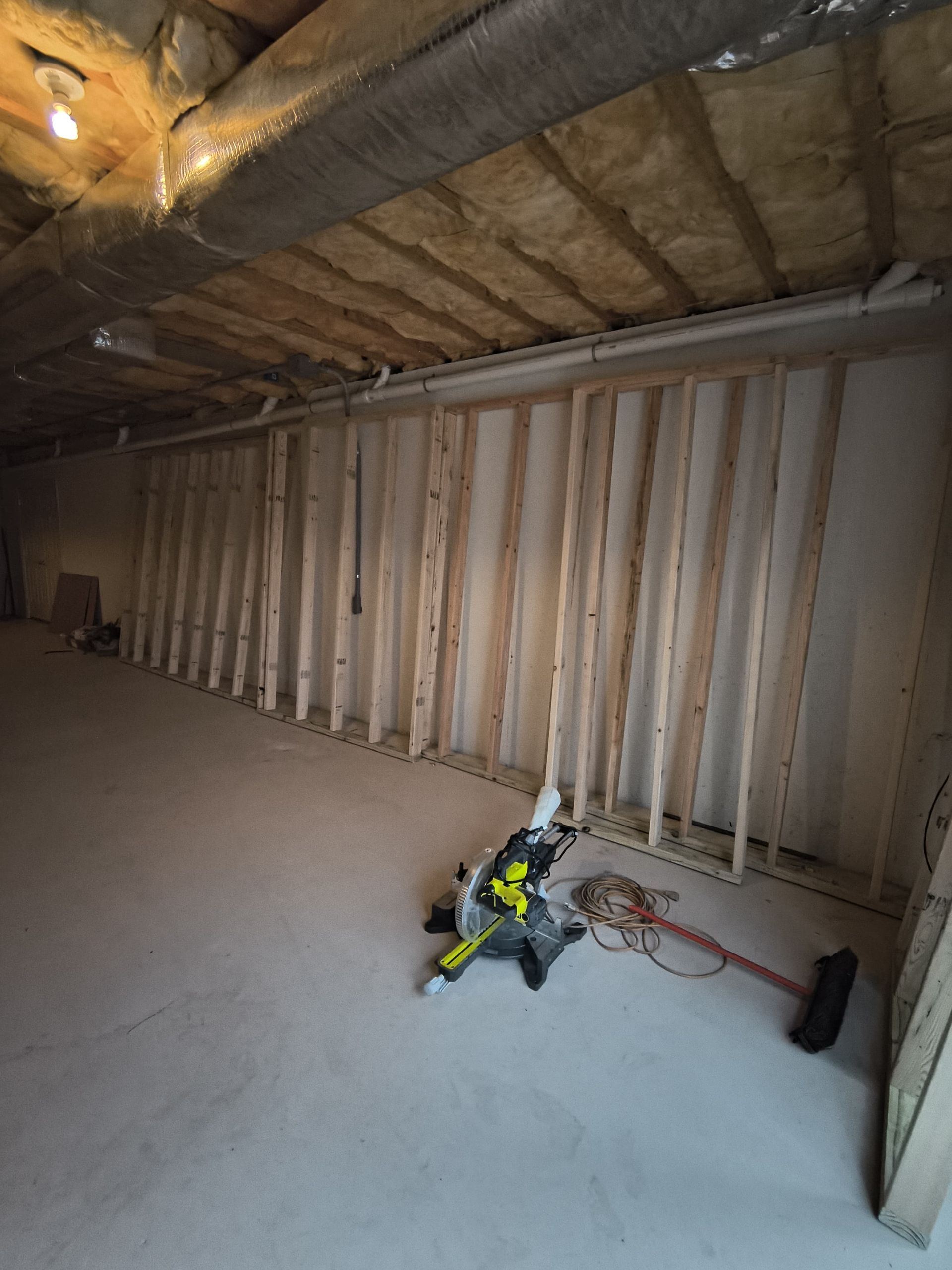 Interior view of unfinished construction. Wooden studs frame a wall, saw and tools on concrete floor.