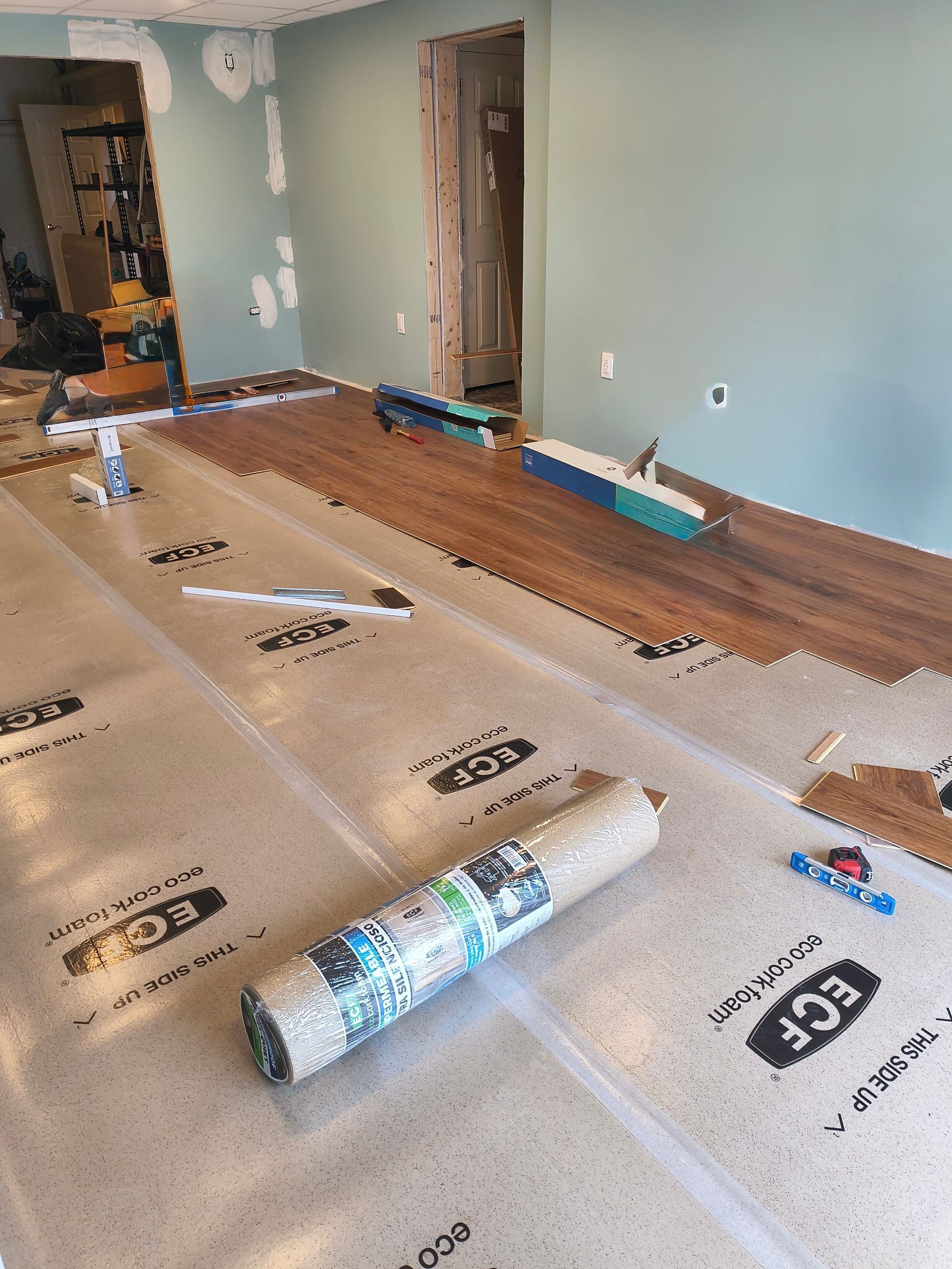 Laminate flooring being installed on a floor with underlayment. The room has teal walls and an open doorway.