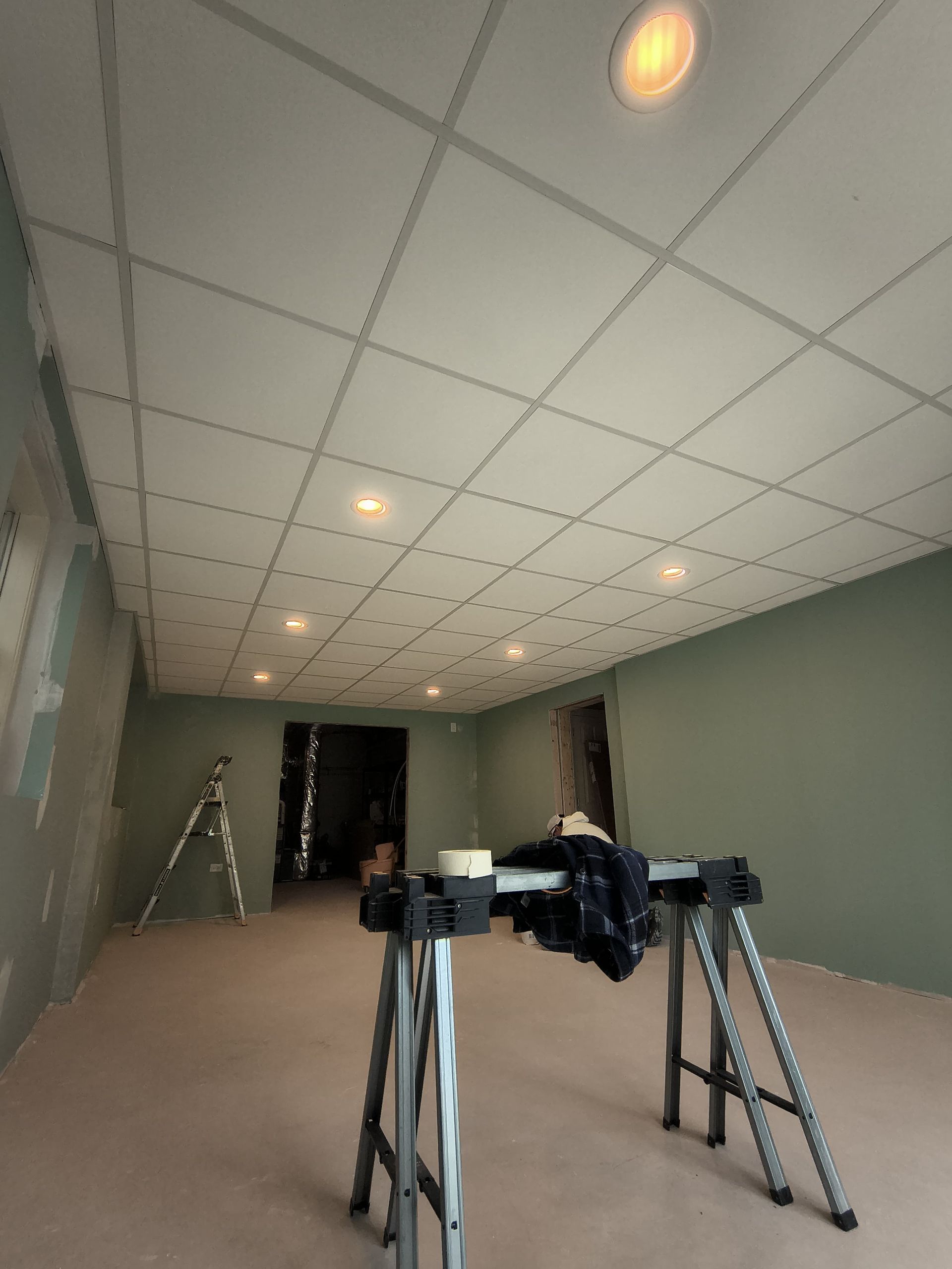 Basement under renovation with a grid ceiling, recessed lights, drywall, and sawhorses.