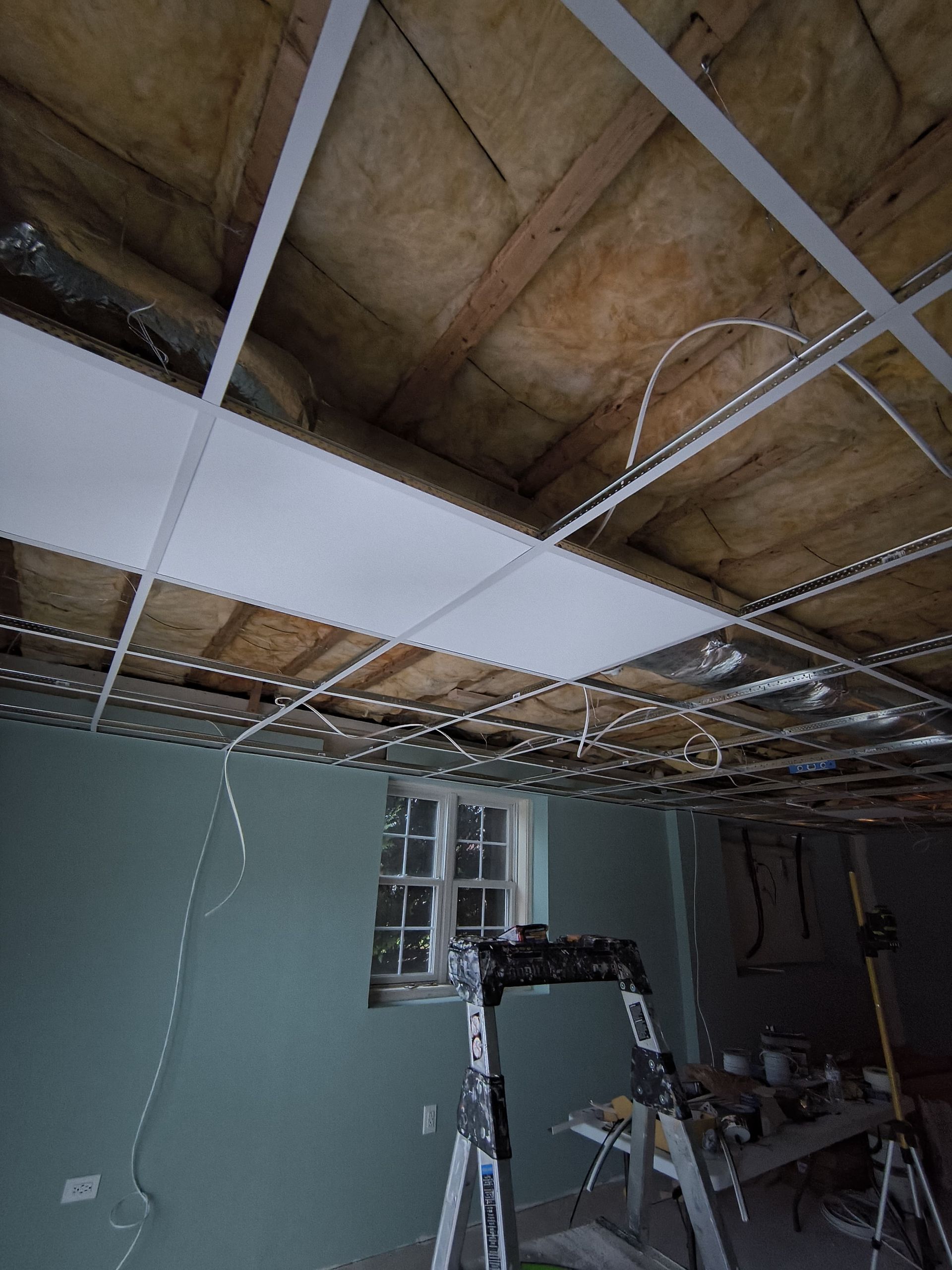 Ceiling construction in a room; grid system with insulation and some panels installed. Ladder and tools are present.