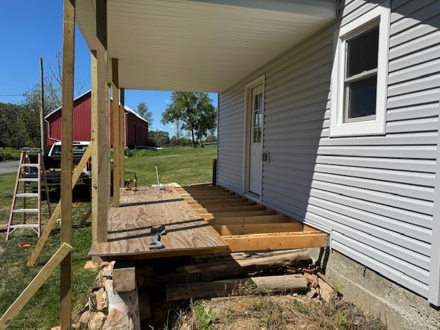 Porch under construction with wooden frame and siding, beside a house with white door and window.