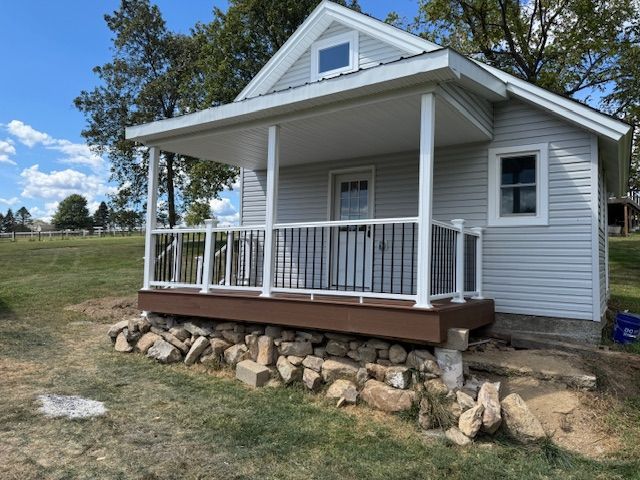 Small gray house with a porch. Brown deck, white railing, and stone foundation. Green grass and blue sky.