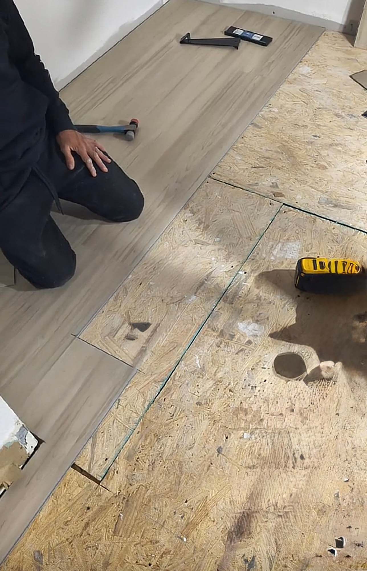 Person installing wood-look flooring. Partially installed planks, tools on floor. Interior setting, tan and black.