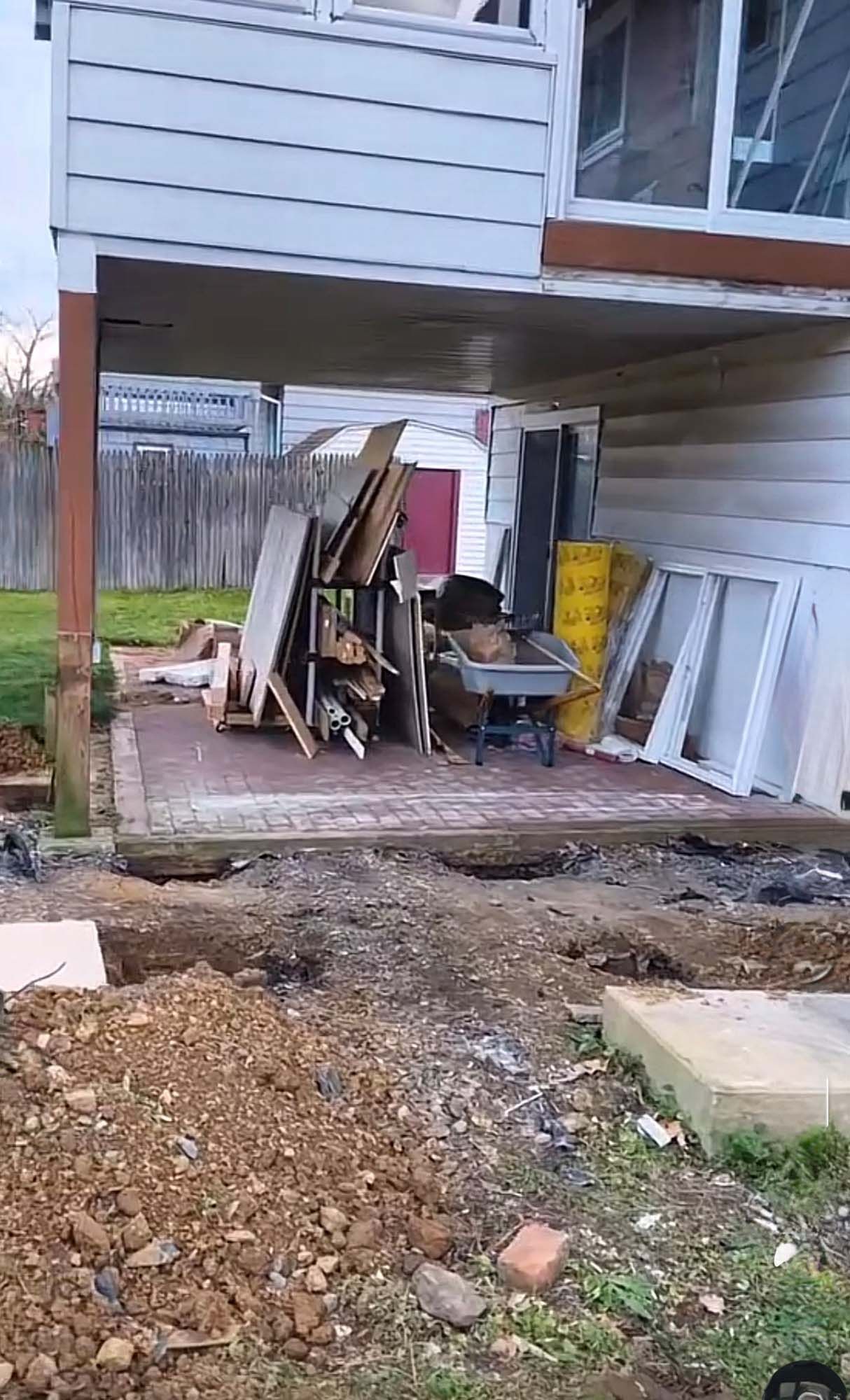Backyard patio under a house with construction materials; dirt and debris in foreground.