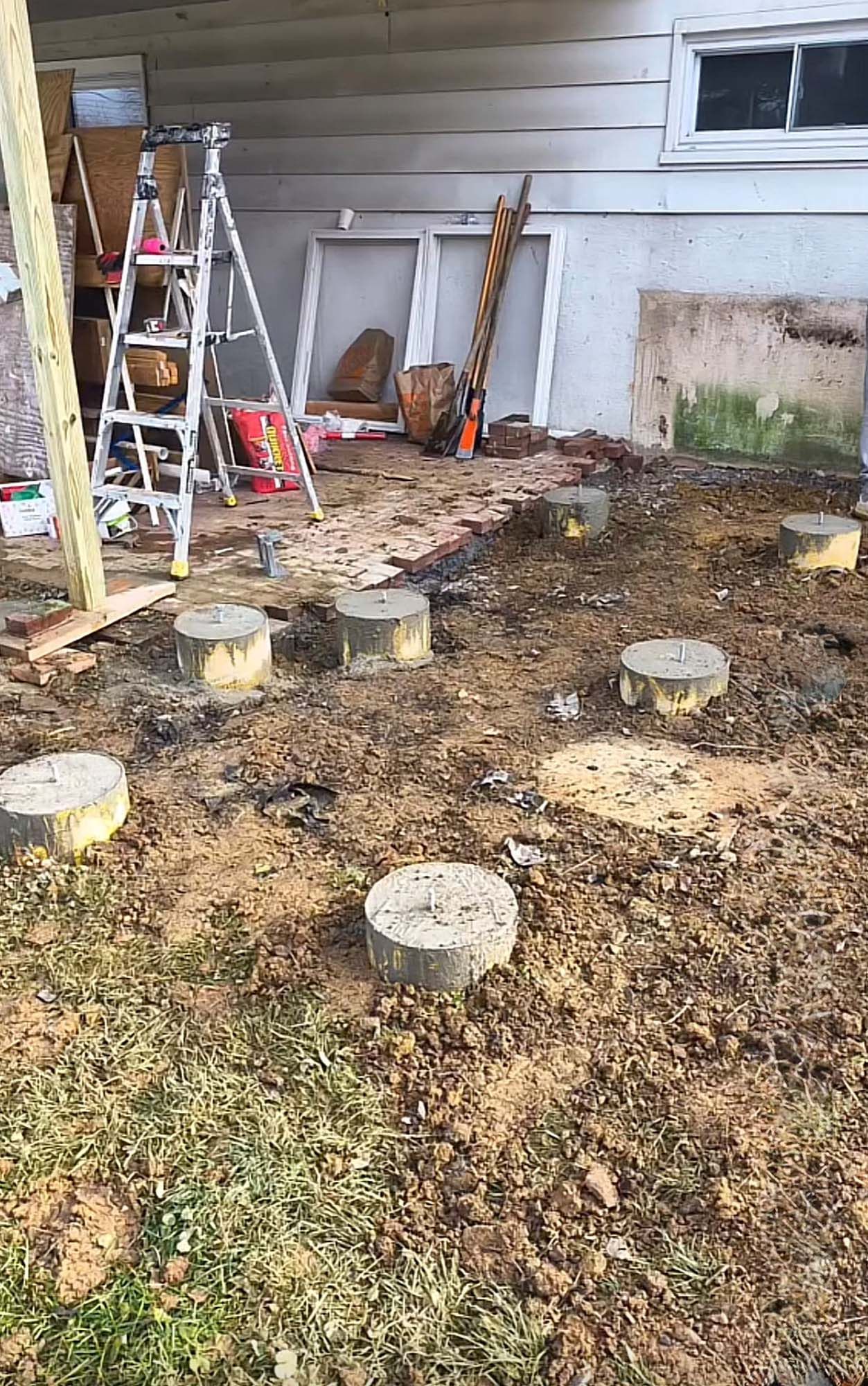Construction site with concrete footings in dirt, ladder, lumber, and house.