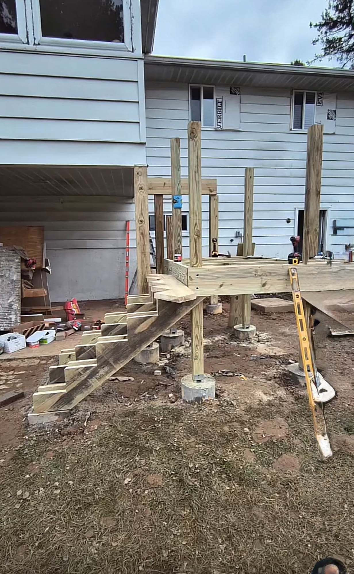 Deck under construction with wooden frame, stairs, and concrete footings near a two-story house.