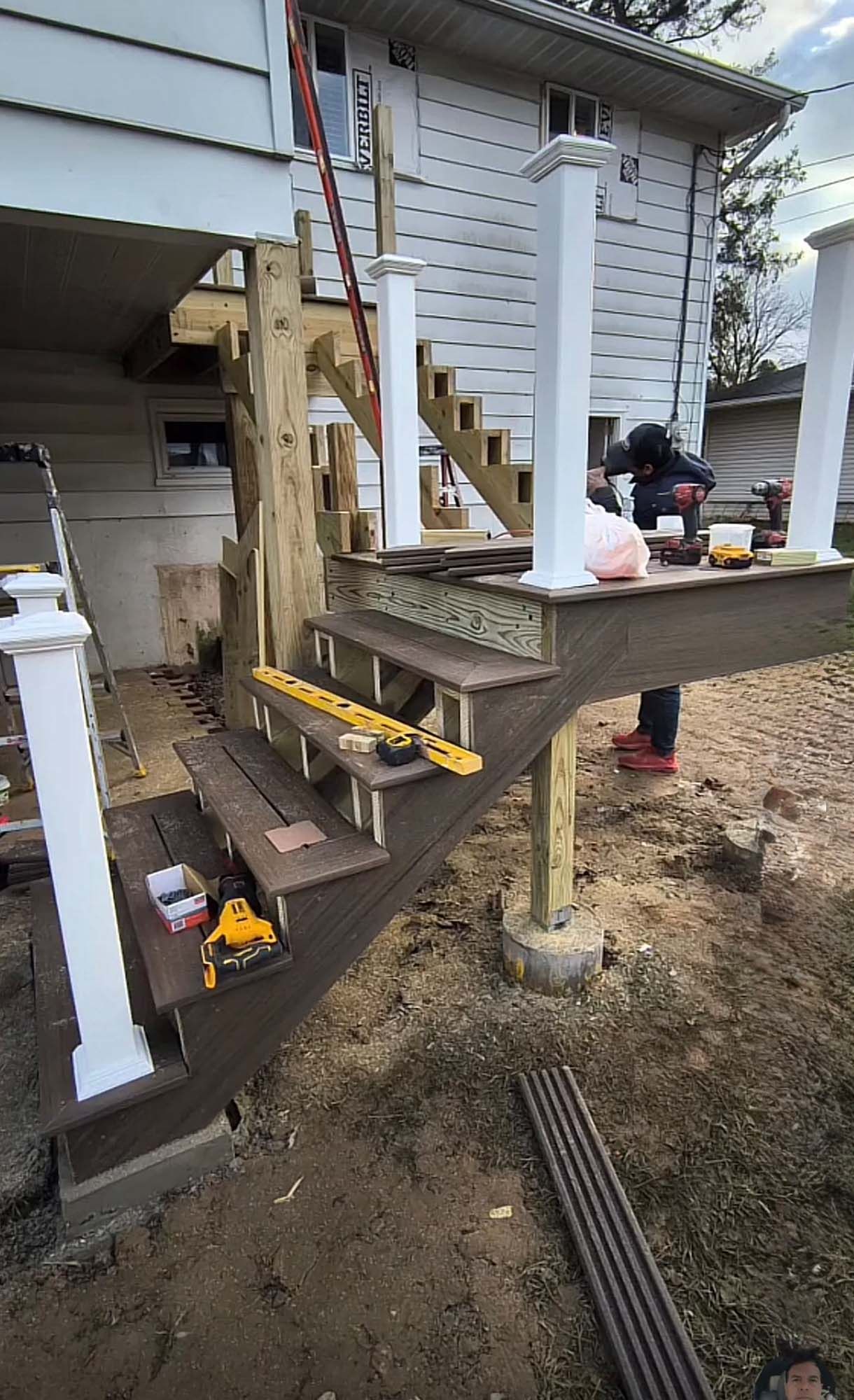 Construction of an outdoor stairway and deck; workers and tools visible. Brown composite and wood, white pillars.