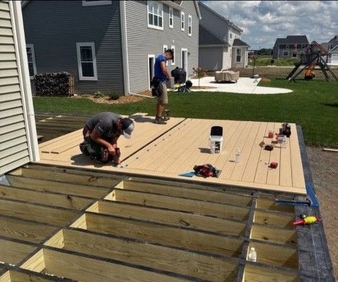 Two workers building a deck; one kneeling, the other standing. Beige deck boards are being installed.