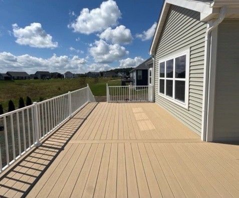Beige deck with white railing next to a gray house, sunny day, blue sky with clouds.