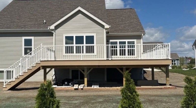 Back of a house with a deck and white railing. Tan deck with stairs, light siding, and green shrubs.