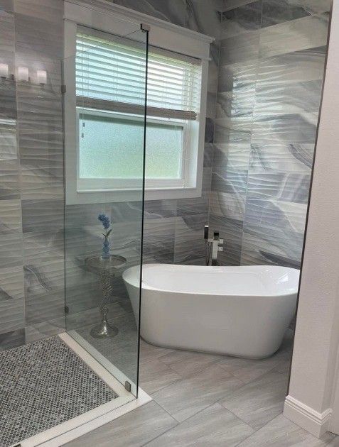 Modern bathroom with a bathtub next to a window and a glass-enclosed shower. Gray and white tiles.
