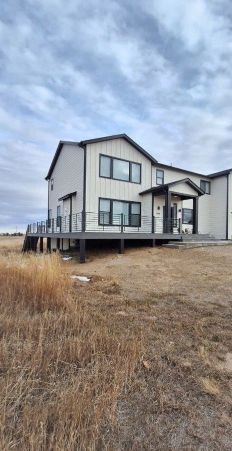 Two-story white house with dark trim and a raised deck on a grassy field under a cloudy sky.