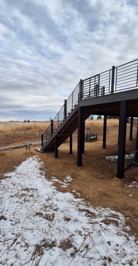 Wooden deck and stairs with black railing in a grassy outdoor setting, patches of snow visible, cloudy sky.