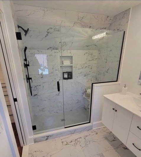 Modern bathroom with a marble-tiled shower behind glass doors. Black shower fixtures and white vanity are visible.