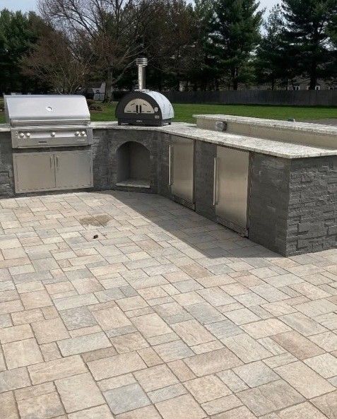 Outdoor kitchen with grill, pizza oven, and counter built into a stone structure on a paved patio.