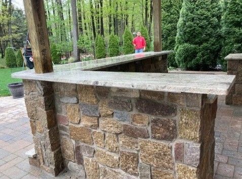 Stone outdoor bar with granite countertop, person in red shirt in background.