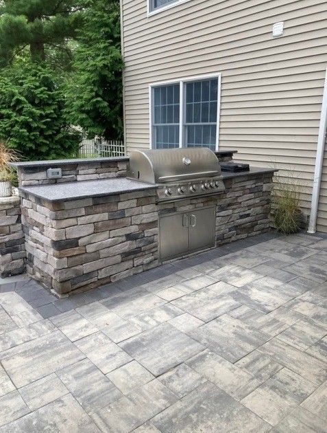 Outdoor kitchen with built-in grill, stone facade, and concrete paver patio next to a house with beige siding.