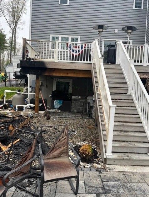 Burnt deck and patio with destroyed furniture under a two-story home, smoke damage is visible.