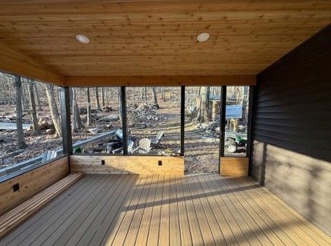 Screened-in porch with wood ceiling and floor, large windows overlooking a forest. Dark siding on the right.