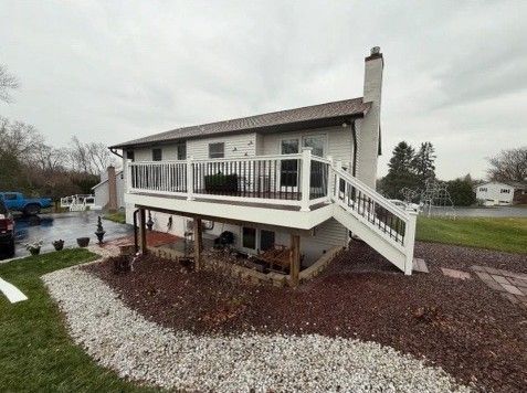 White house with a raised deck, stairs, and a dark railing, surrounded by a red mulch landscaping and a stone path.
