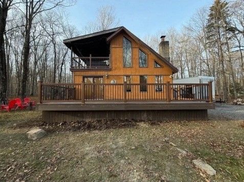 Wooden cabin with a deck, surrounded by trees. Brown exterior, tall windows, and a chimney.