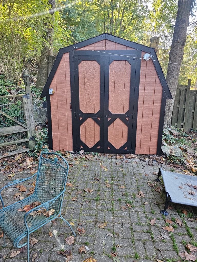 Brown and black shed with double doors, brick patio, and green metal chair outdoors.