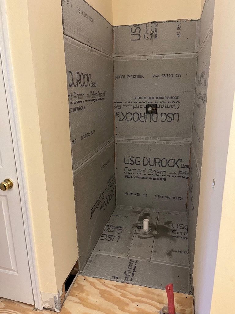 A shower stall under construction; gray cement board walls, plumbing visible, open doorway on the left.