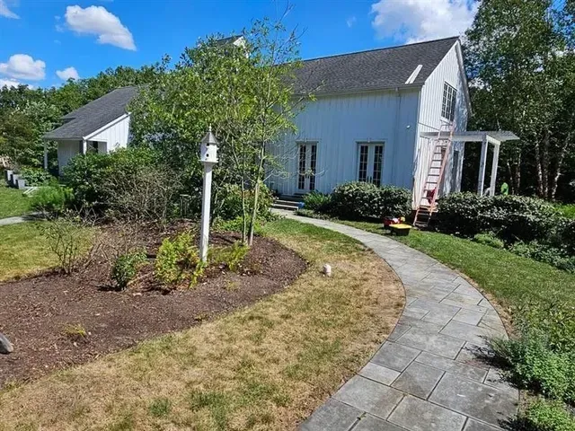 White house with a gray stone pathway, birdhouse, and landscaping on a sunny day.