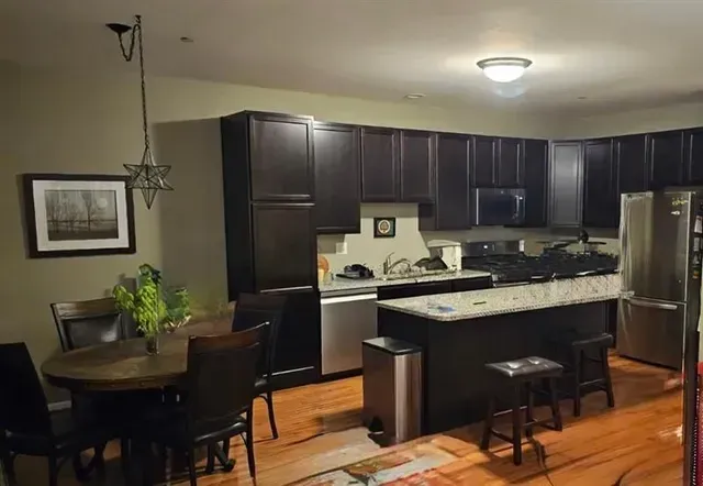 Kitchen with dark brown cabinets, island, and dining table. Stainless steel appliances and hardwood floors.