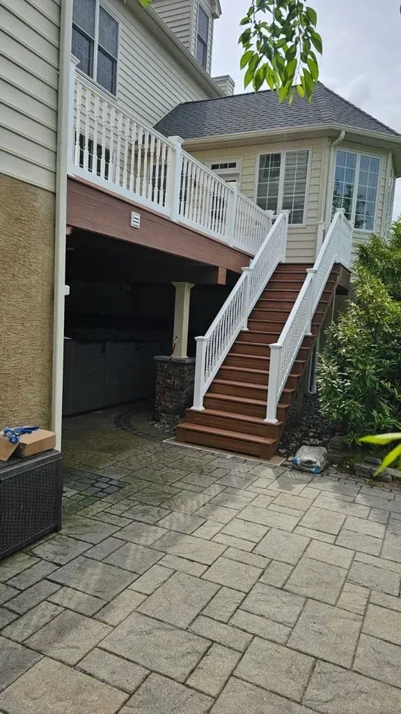 A wooden deck and staircase lead to a house with a stone patio and landscaping.