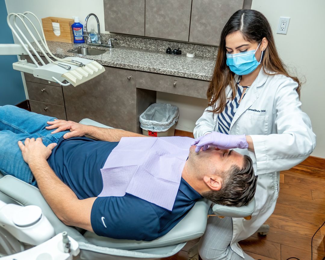 A male patient is reclined on a dental chair while Dr. Kavneet Bindra examines his teeth